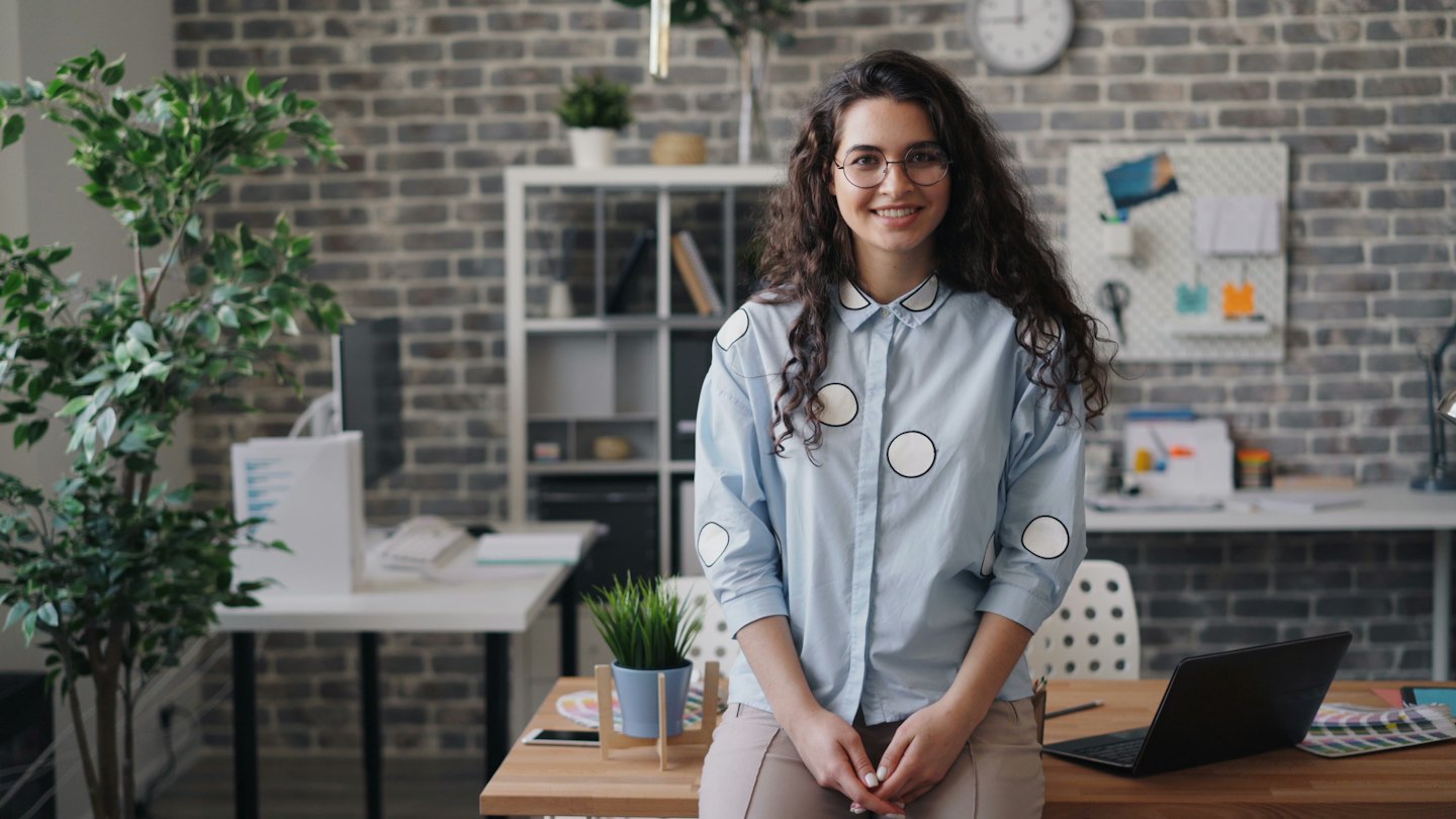 Young woman in polka-dot shirt smiles at desk with laptop.